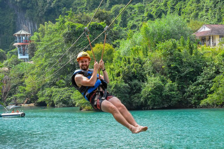 A man zip lining over turquoise water to dark cave in Phong Nha, Vietnam