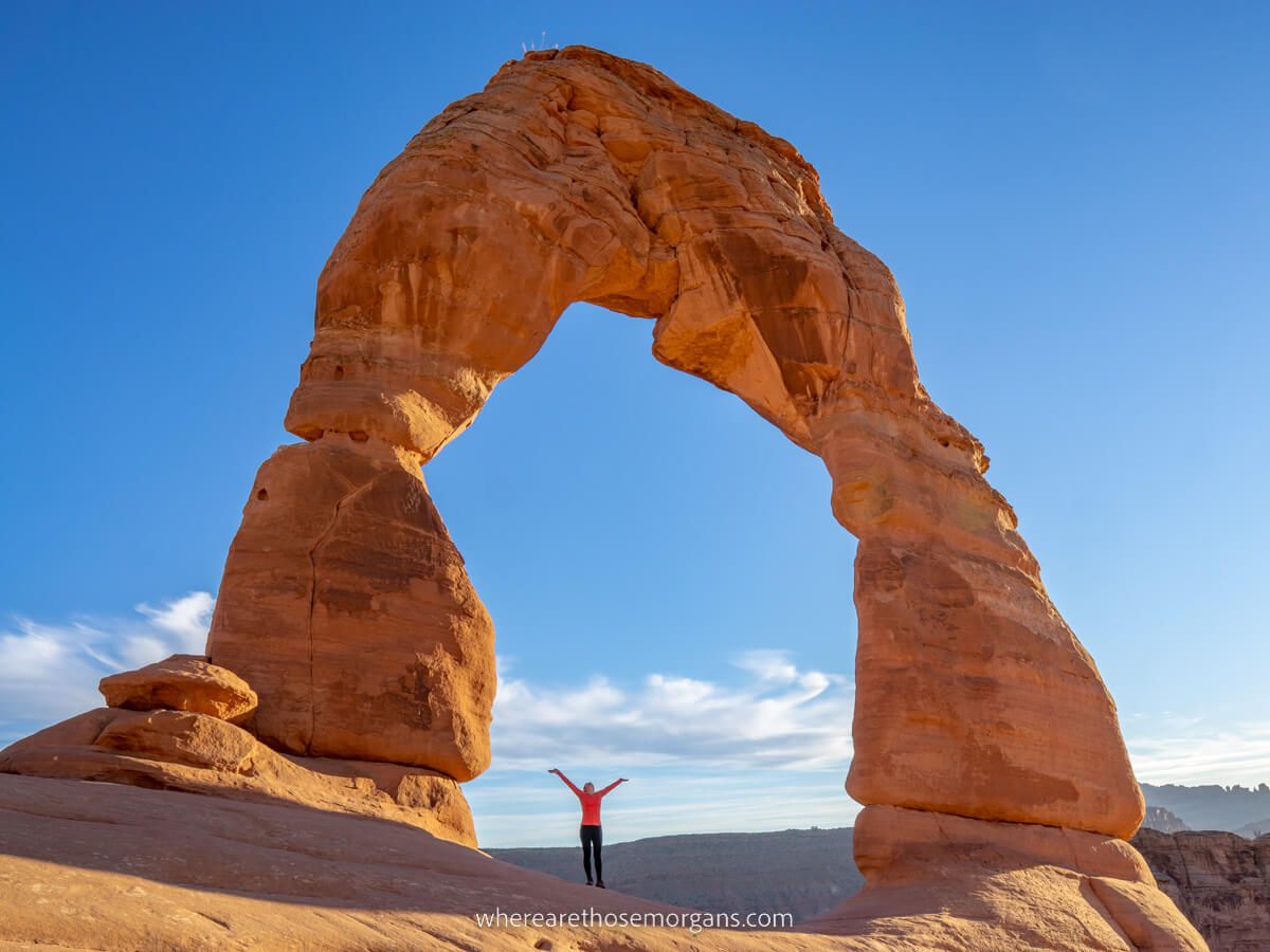 Woman looking tiny under the gigantic Delicate Arch formation on a day trip to Arches and Canyonlands national parks in Utah