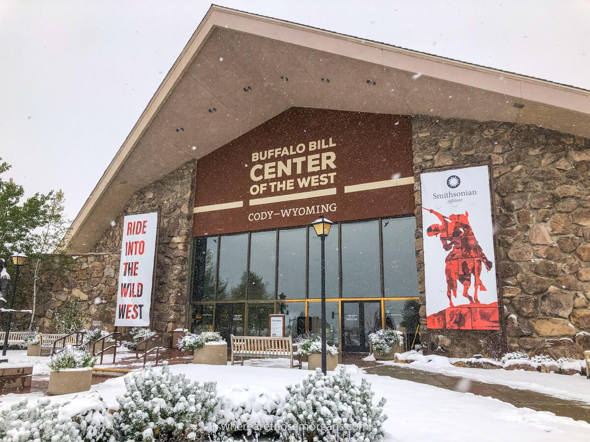 Exterior photo of a building made of large stones with a triangular roof and glass windows at the front with Buffalo Bill Center of the West Cody Wyoming printed above on a snowy day