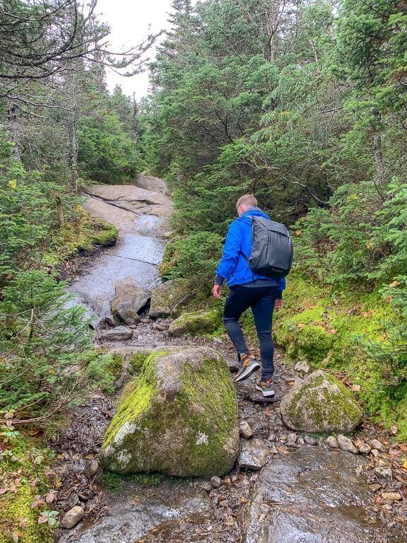 Man on trail with boulders and trees in adirondacks new york with blue coat and peak design backpack and adidas terrex free hikers