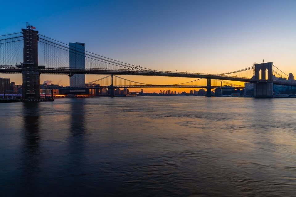 Beautiful photograph of Brooklyn Bridge and Manhattan Bridge from Pier 17 in New York City at dawn just before sunrise orange sky and smooth river