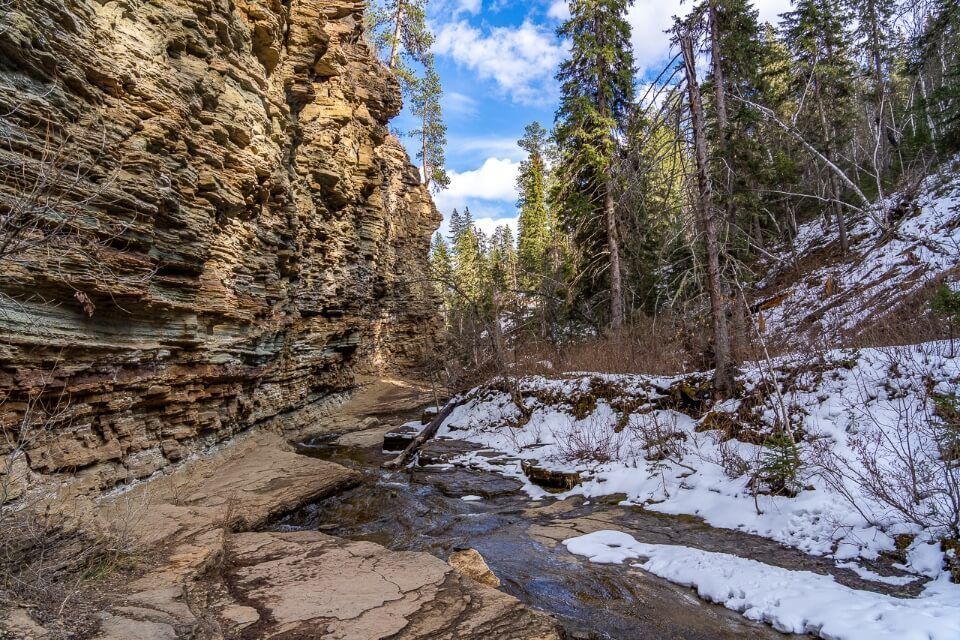 Narrow creek with snow on the ground and a tall cliff wall to one side