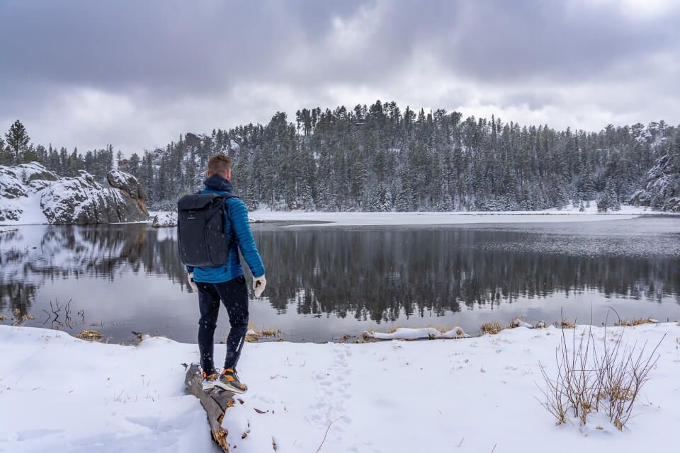 Hiker standing on log covered in snow in front of a lake surrounded by ice