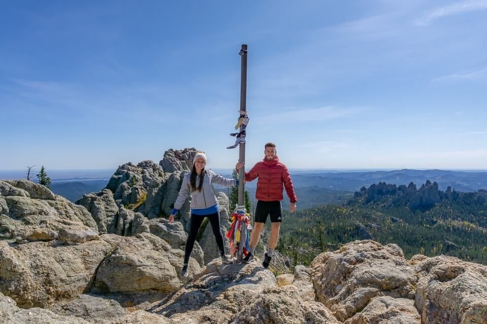 Mark and Kristen from Where Are Those Morgans holding onto a flagpole at the summit of Black Elk Peak hiking trail near Mount Rushmore in South Dakota