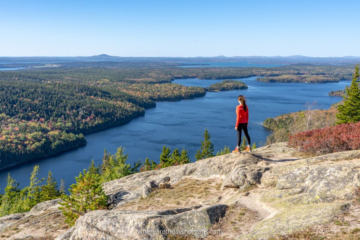 Hiker looking out at lakes and rolling hills covered in trees from a rocky ledge in the fall