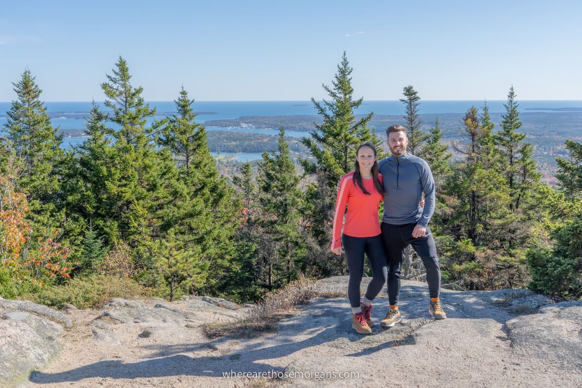 Couple standing together in hiking clothes at the top of a mountain peak with trees and distant views over the ocean behind