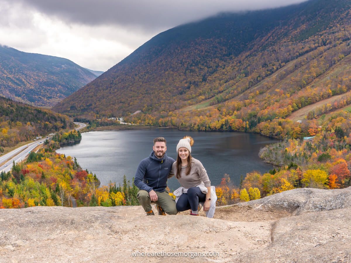 Two hikers crouched next to each other on a rocky outcrop with views over a lake and rolling hills covered in vibrant fall foliage colors on a cloudy day at Artists Bluff in New Hampshire