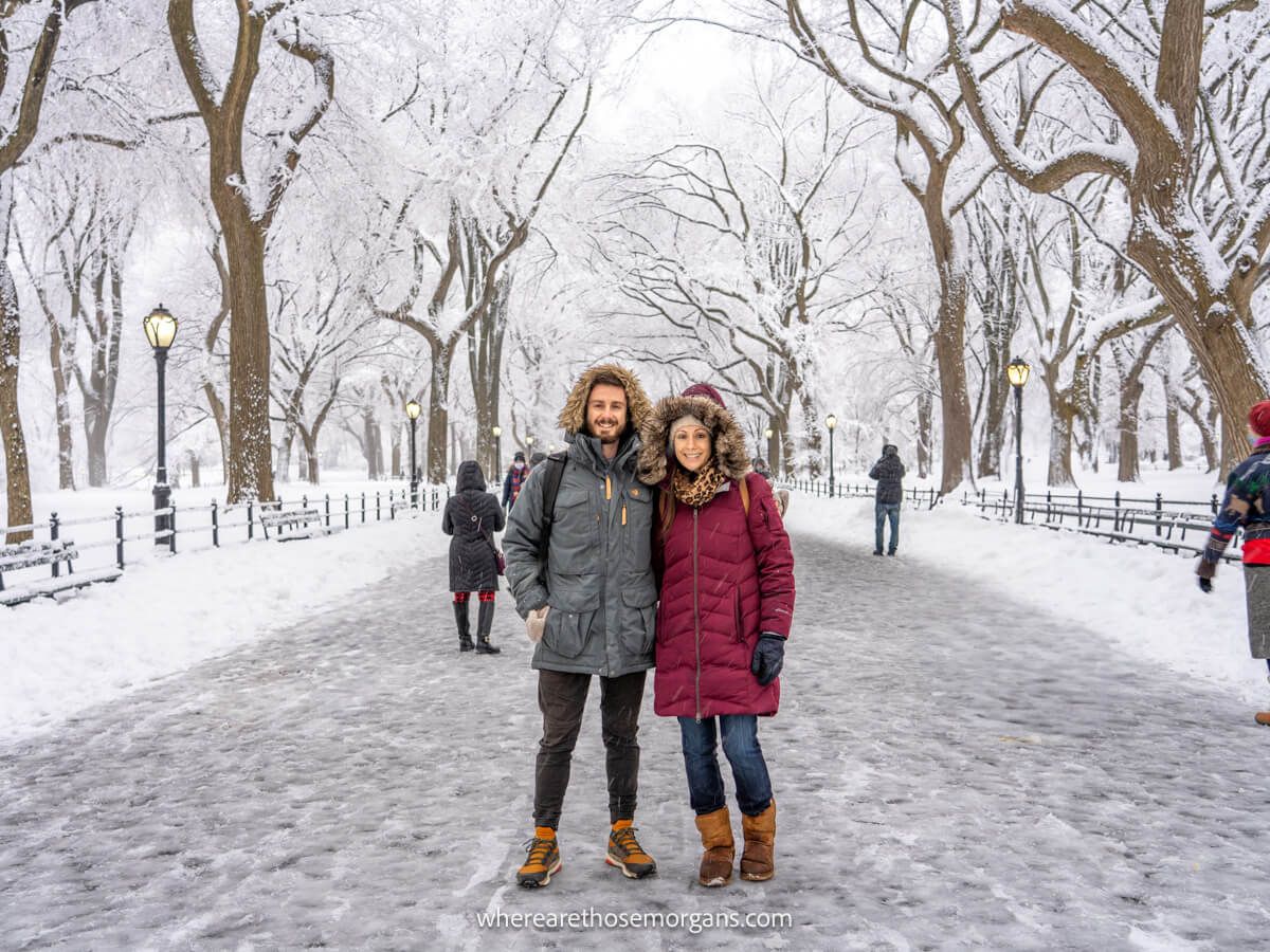 Photo of a couple standing together in heavy winter coats on a snowy path surrounded by bare trees covered in snow and more snow on the ground in Central Park NYC