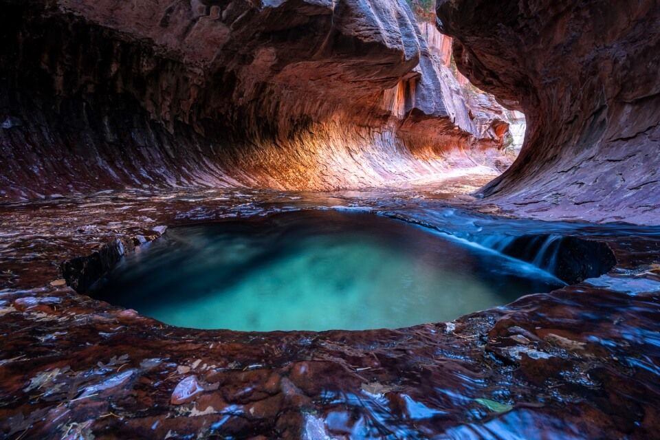 Small turquoise colored pool in a tunnel like cave with orange light glowing on a hike in Zion