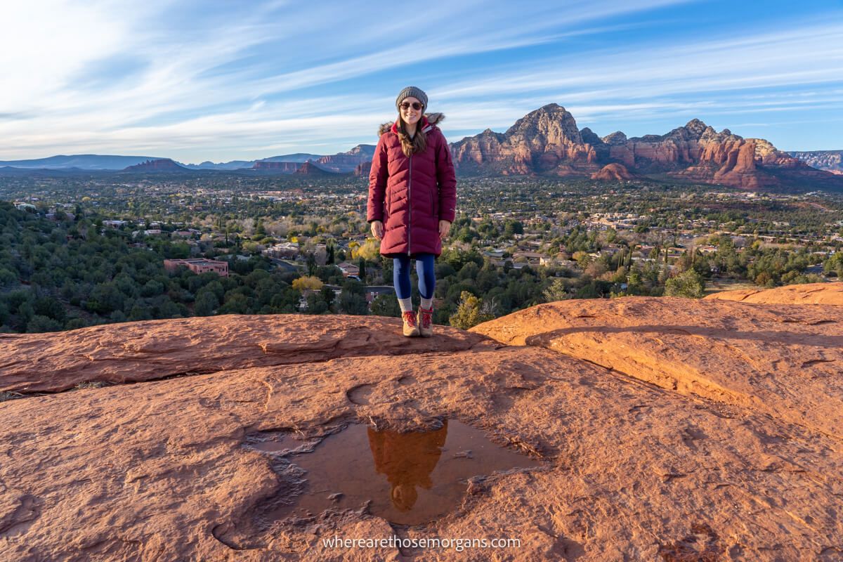 Hiker in thick red coat standing on a red rocky mound with reflection in a small puddle and far reaching views over a valley and towering red rocks in the distance