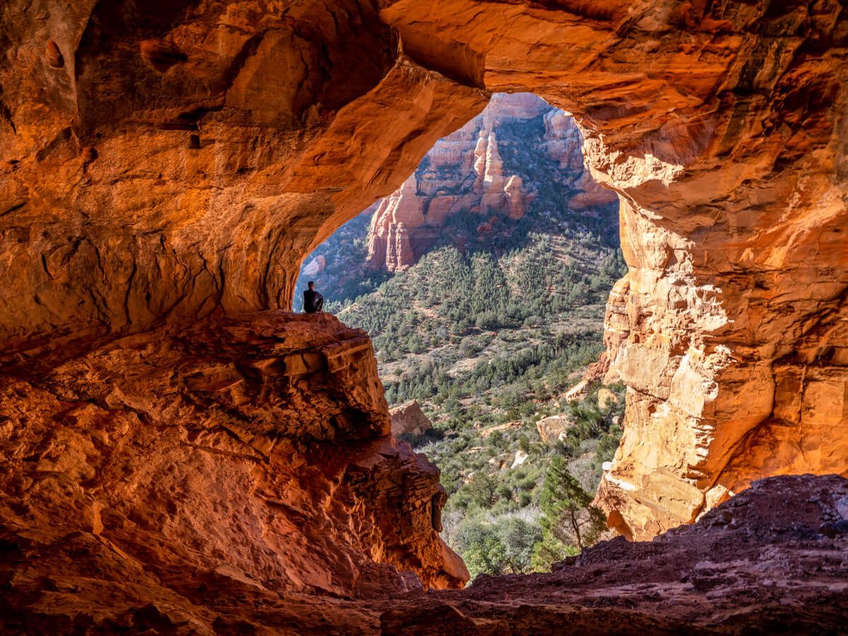 Photo of a hiker sat on the edge of a ledge looking out of a giant cave entrance to the valley below