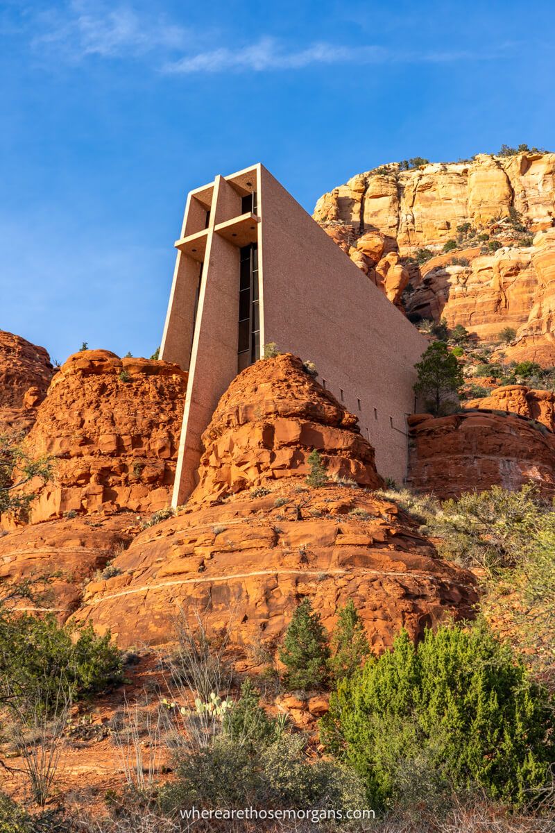 Looking up at a church shaped like a cross built into a red rock cliff on a clear day