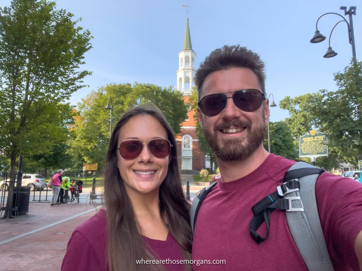 Couple in maroon shirts standing together taking a selfie with a church behind in Burlington Vermont