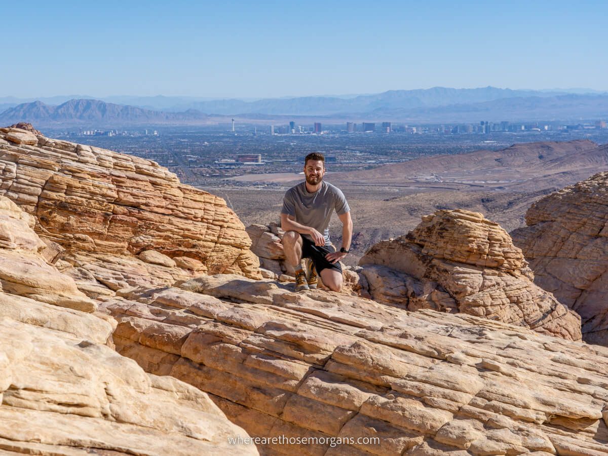 Hiker crouched at summit of Calico Tanks one of the best hikes in Red Rock Canyon with Las Vegas strip in background