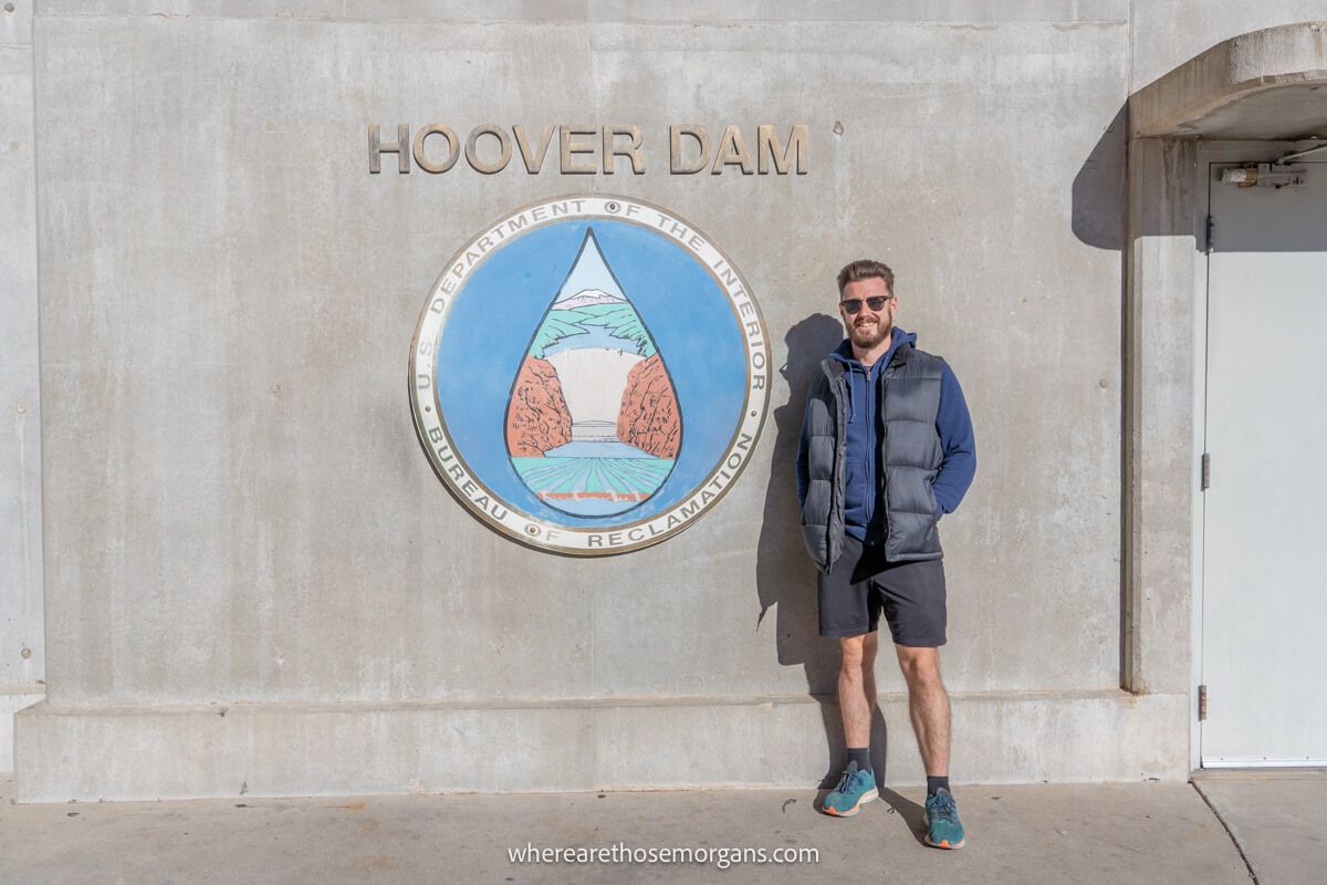 Tourist in shorts and vest stood next to the Hoover Dam sign on a concrete wall