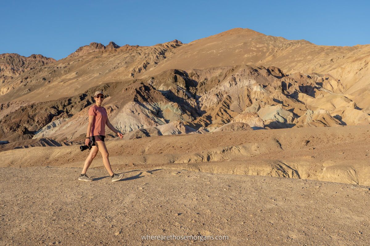 Hiker holding a camera and walking on a dirt mound with more colorful mounds in the background on a sunny day in Death Valley National Park