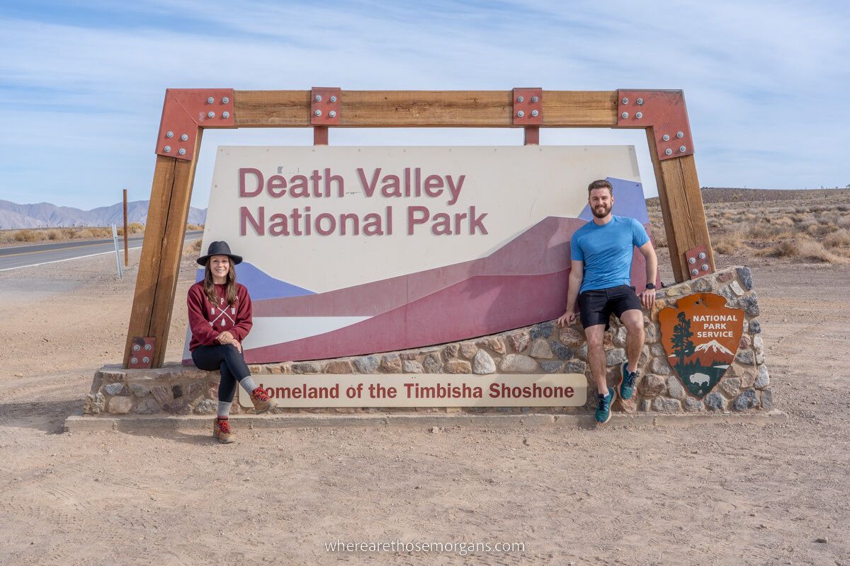 Couple sat on either side of the Death Valley National Park welcome sign on a warm day with light clouds