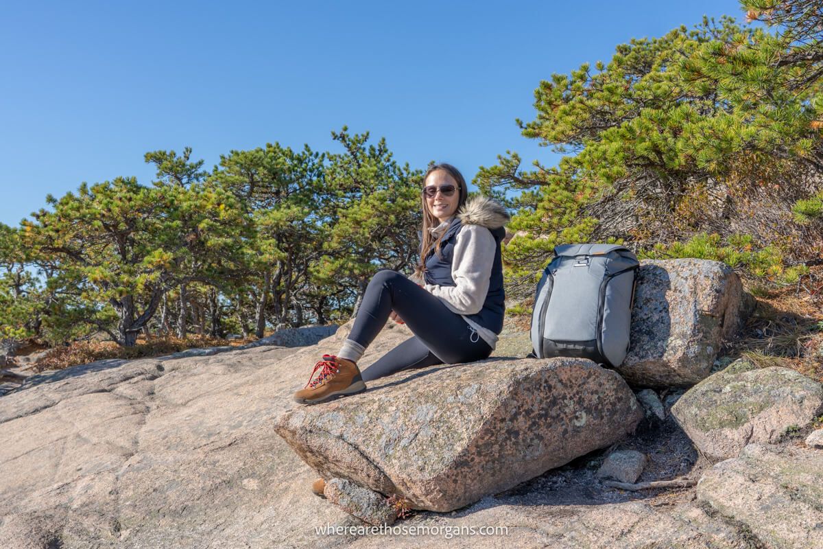 Hiker with boots, fleece and sunglasses sat on a boulder with a grey backpack with trees and a blue sky behind