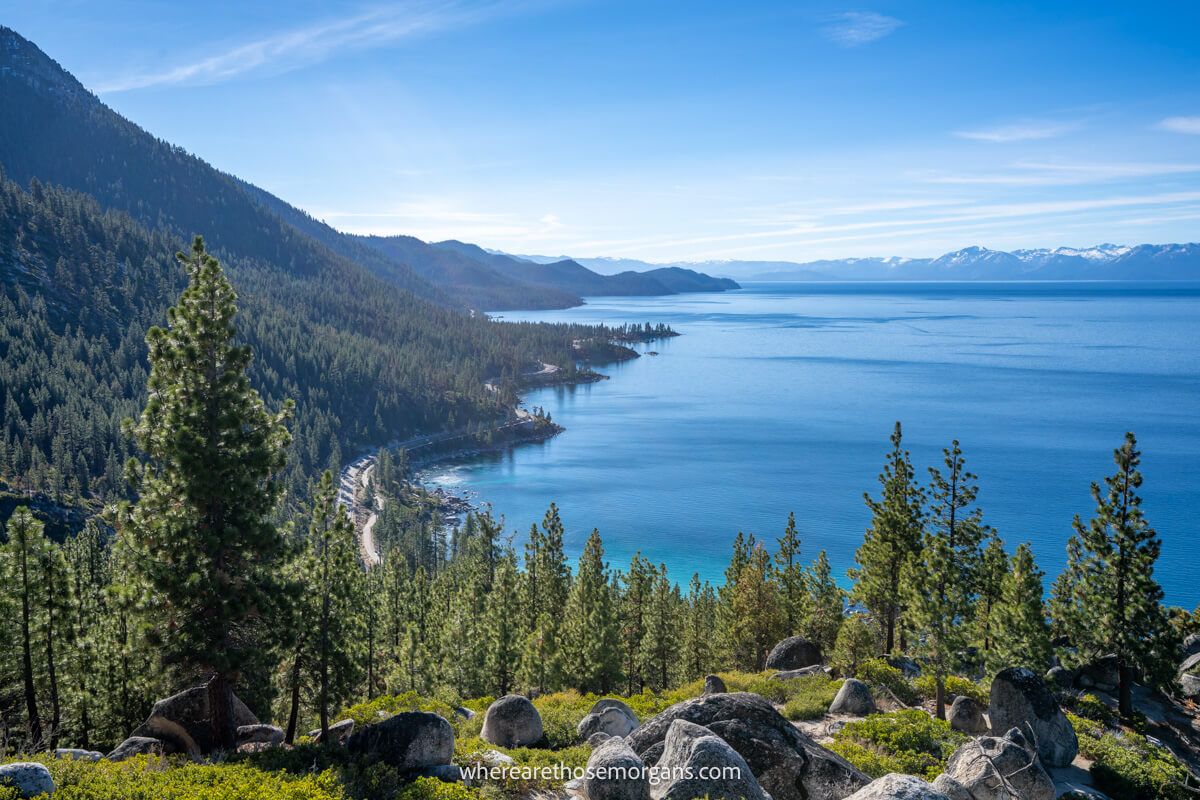 Photo of Lake Tahoe's eastern shoreline from Monkey Rock with trees and rocks in the foreground, steep slopes in the middle in snow capped mountains in the distance