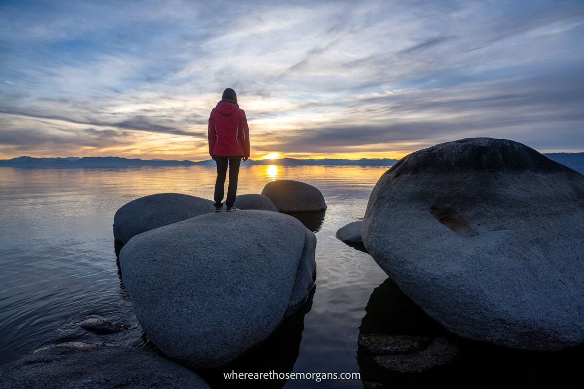 Hiker in light coat standing on a smooth large boulder in a lake with the sun setting on distant mountains