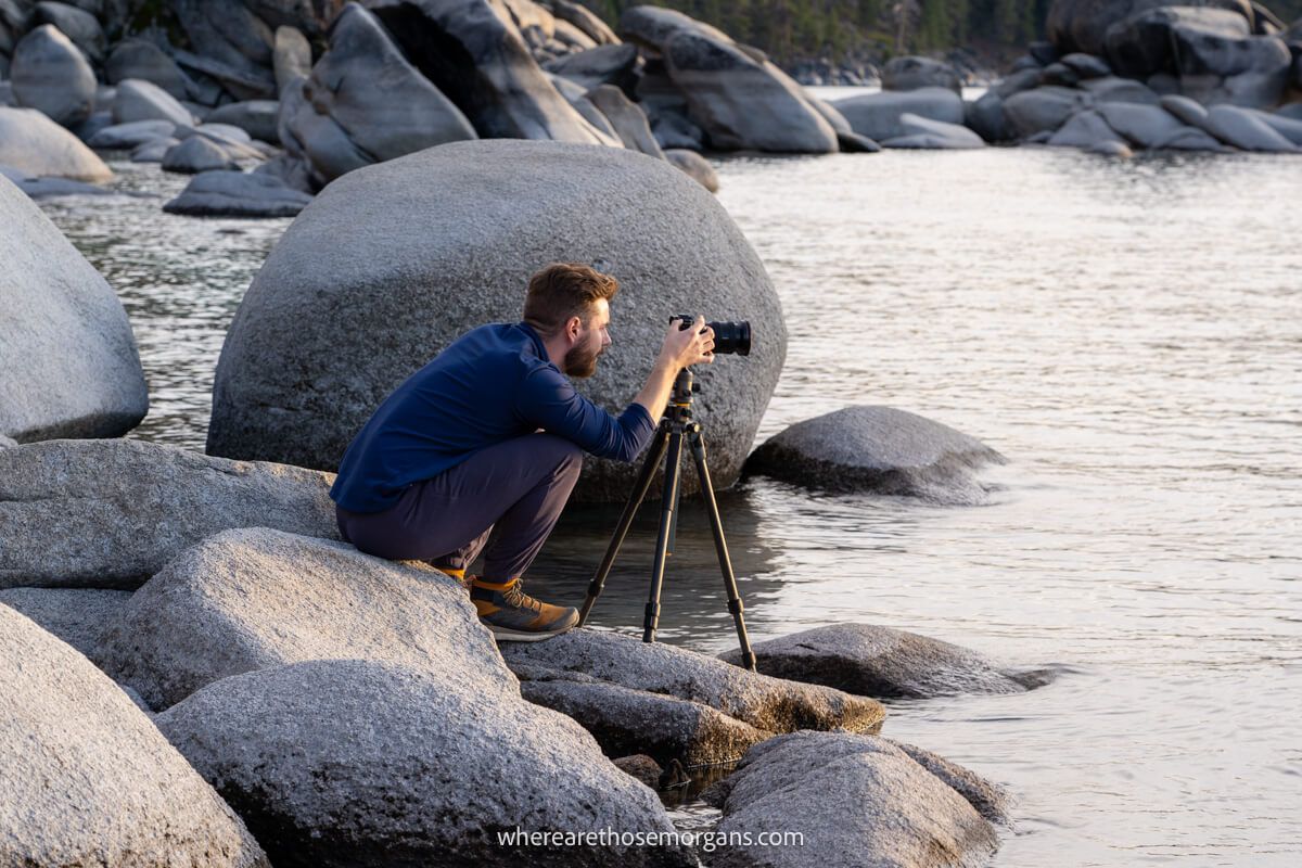 Photographer crouching on a rock next to boulders and water with camera on a tripod