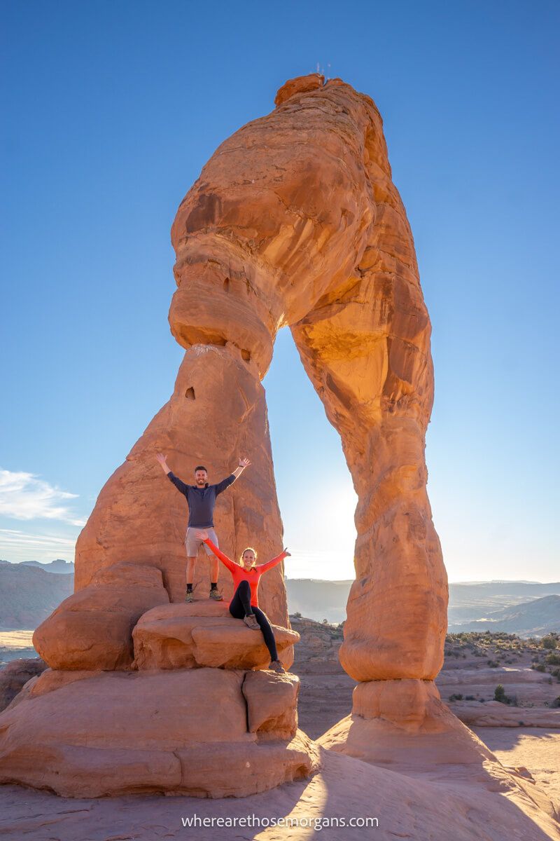 Hikers sat and stood on a giant sandstone arch with arms in the air