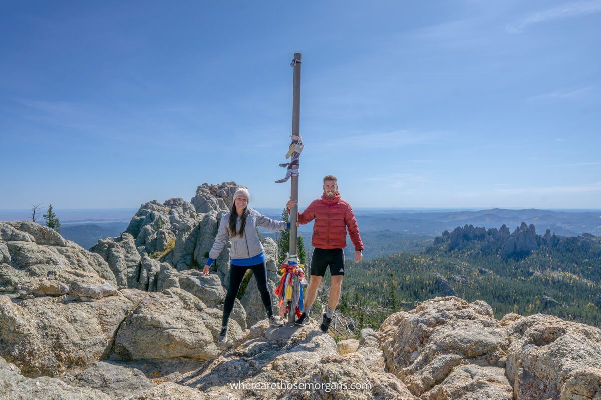Two hikers holding onto a summit flagpole built into boulders with far reaching views over trees on a clear day in Custer State Park, Black Hills
