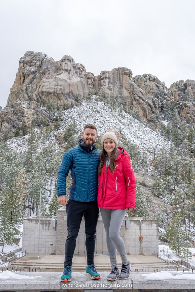 Couple with winter coats stood together for a photo in front of Mount Rushmore on a cold dull and snowy day