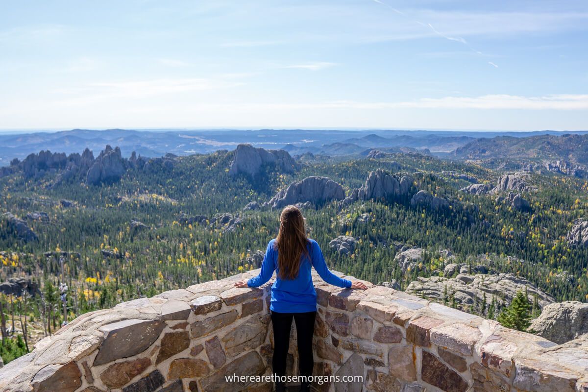 Hiker stood in the corner of a low brick wall looking out at horizon reaching views of trees and granite rocks on a sunny day