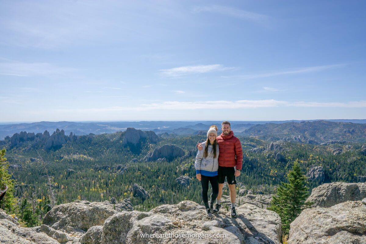 Hiking couple stood together for a photo on granite rocks with distant views over a valley covered in trees and a wide open blue sky