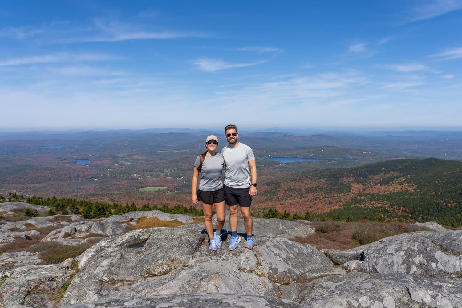 Two hikers stood together for a photo at the summit of a rocky mountain with far reaching views behind