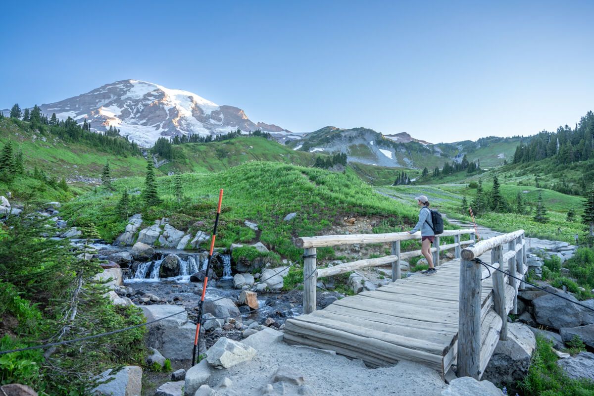 Hiker stood on a wooden bridge looking at green meadows, a stream and a snow capped volcano at dawn