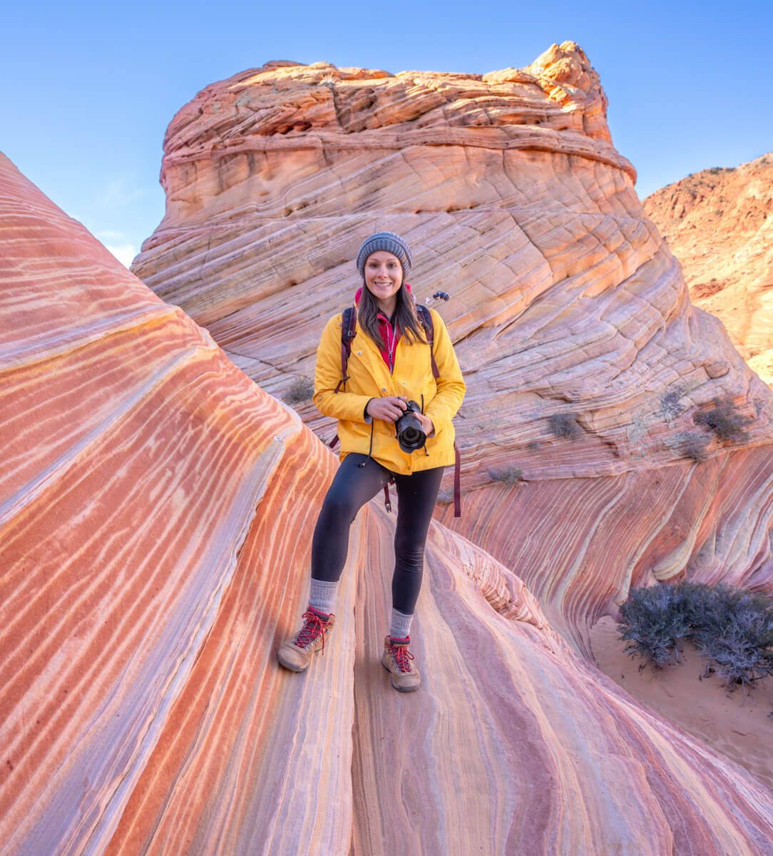 Hiker holding camera in a yellow coat and wooly hat standing on a sloped colorful rock formation with a clear blue sky