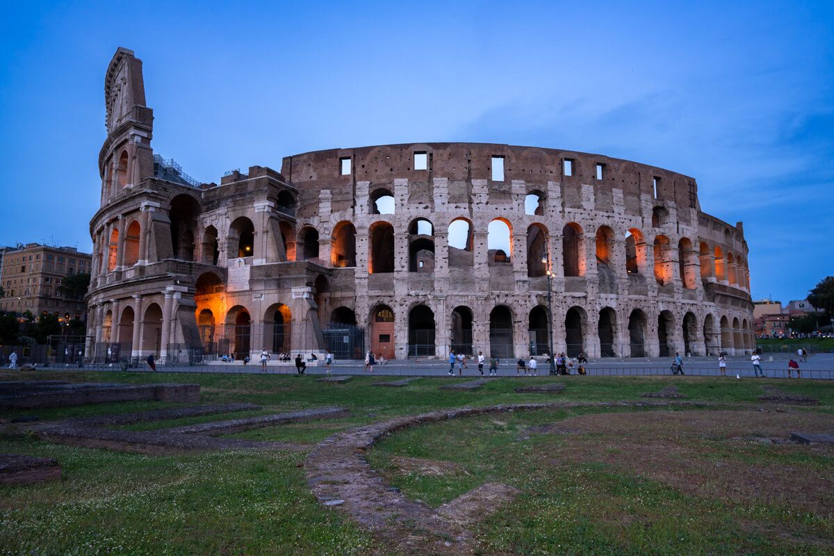 The colosseum of Rome at sunset with orange lights and a deep blue sky