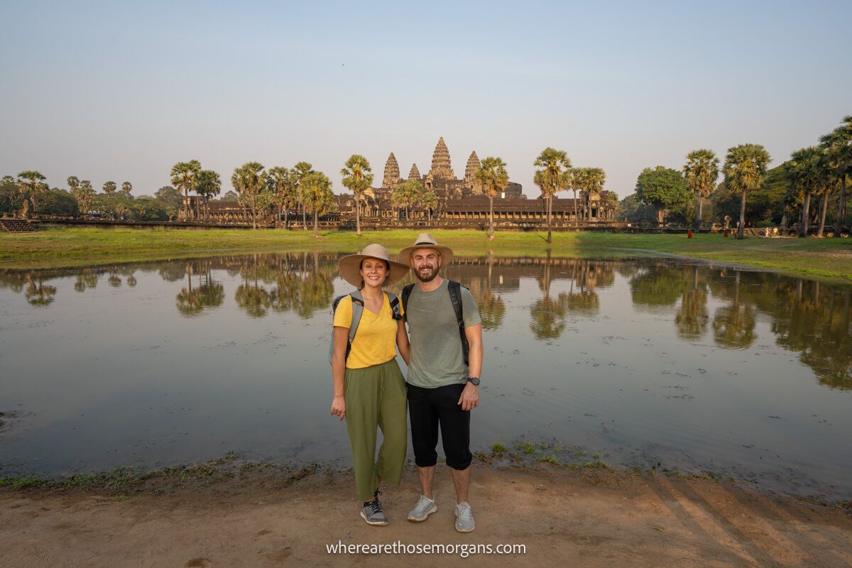 Photo of a couple standing together in sweaty lightweight clothes and hats with a small lake behind and Angkor Wat temple further behind at dusk