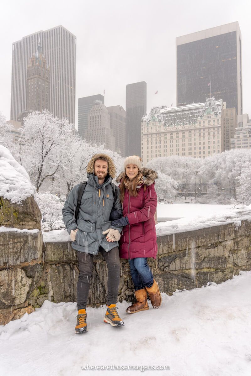Photo of a couple standing together with heavy winter coats on a stone bridge in Central Park NYC on a snowy day