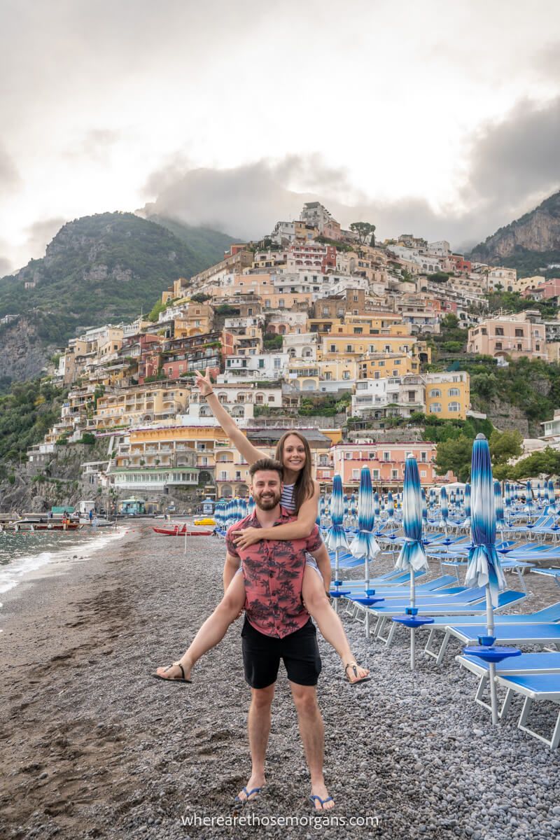 Photo of a couple on a pebble beach looking happy with blue sun loungers to one side and a small town built into a hill behind on a cloudy day