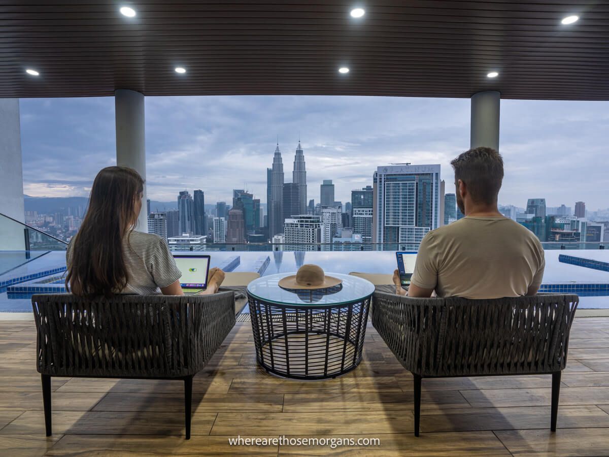Photo of a couple with backs turned sitting on straw sun loungers on a wooden floor with a rooftop pool ahead leading to a city view with skyscrapers