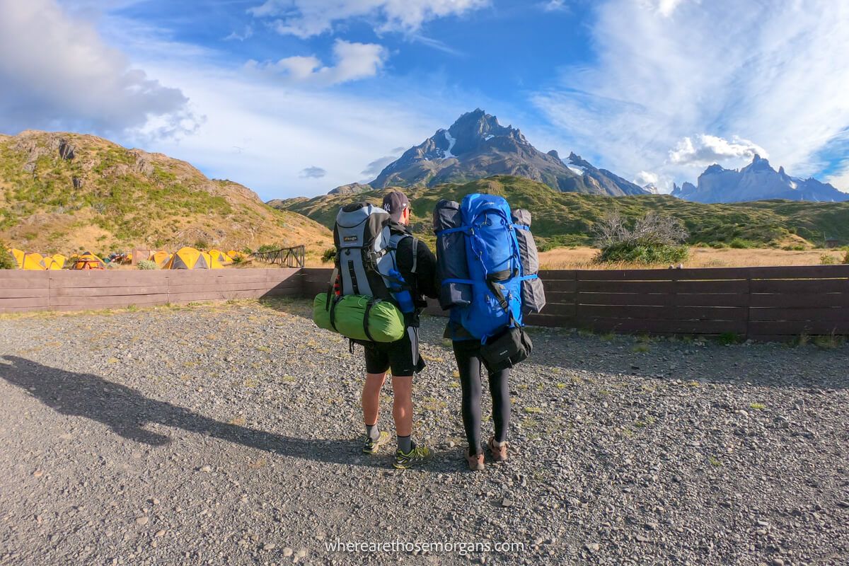 Photo of a couple standing together with backs turned and huge backpacks on in a mountainous landscape with blue sky and light clouds