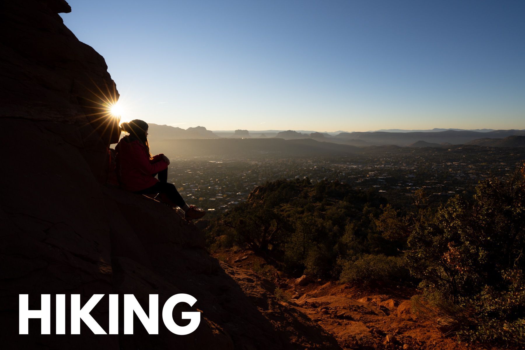 Photo of a hiker sat on a rocky cliff overlooking a wide open view of Sedona at sunrise with the word Hiking overlaid