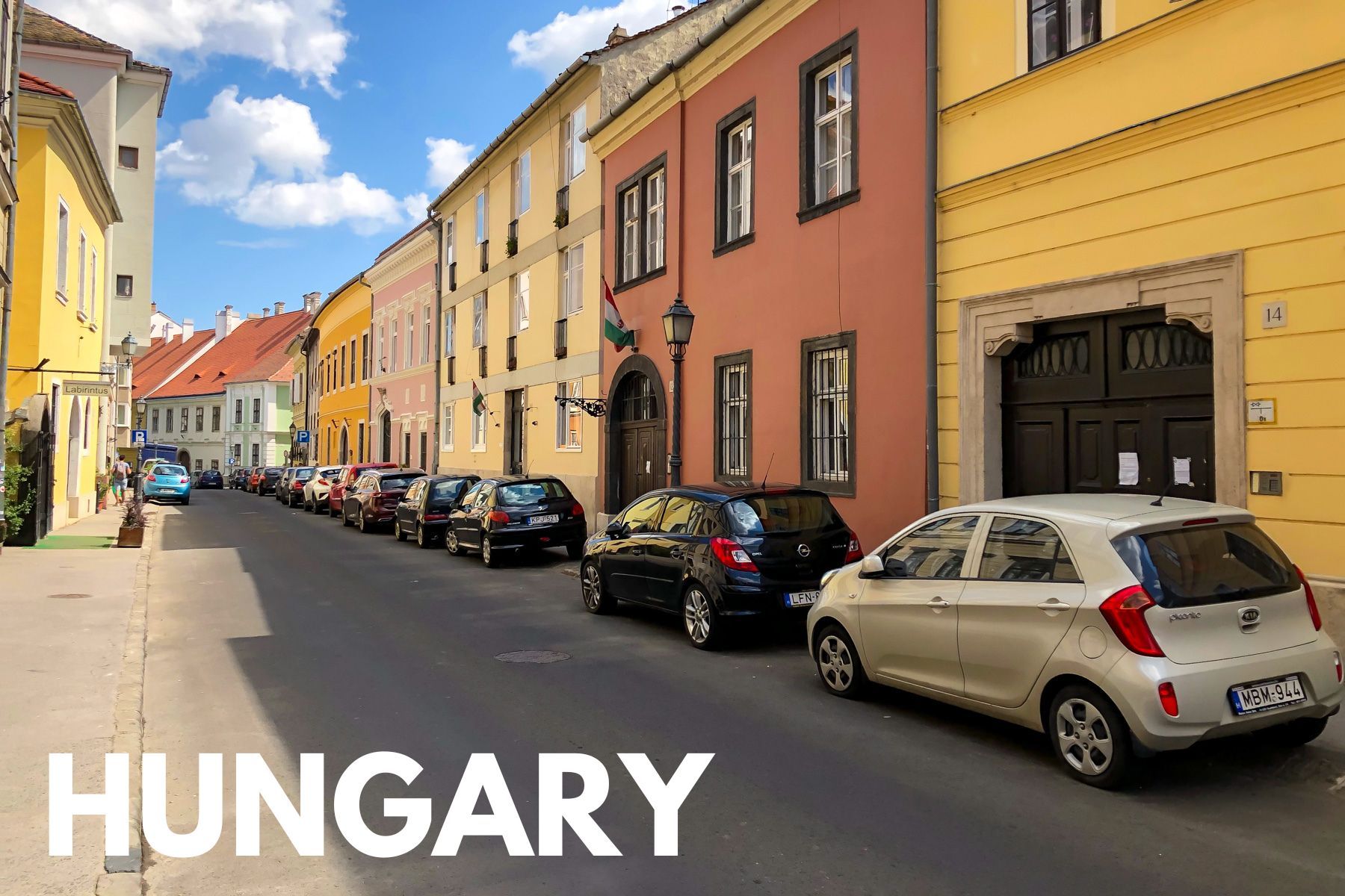 Photo of a row of colorfully painted houses with cars parked outside on a narrow street and the word Hungary overlaid