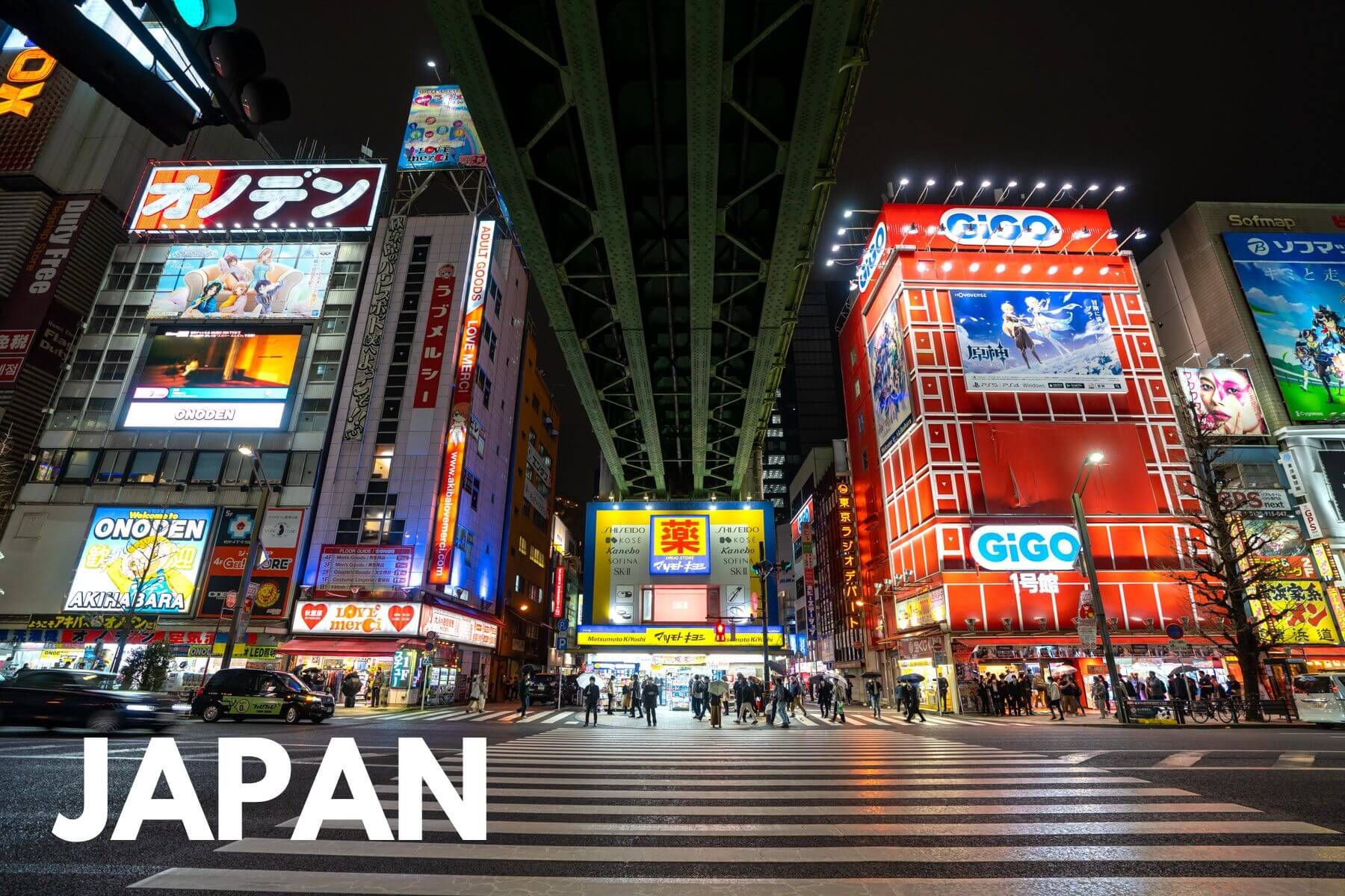 Photo of Akihabara in Japan at night famous video game area with lots of bright lights on tall buildings next to a bridge and a pedestrian crossing with the word Japan written on the photo