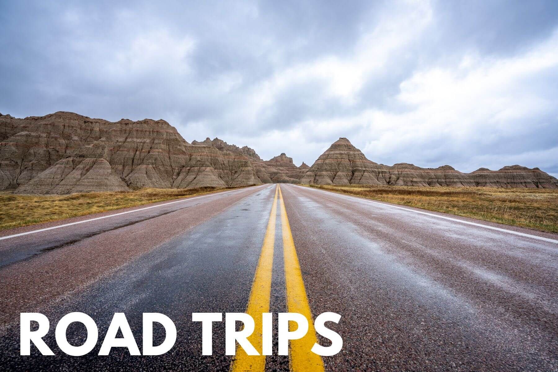 Photo of a road with yellow lines leading into Badlands formations on a cloudy day with the words Road Trips overlaid