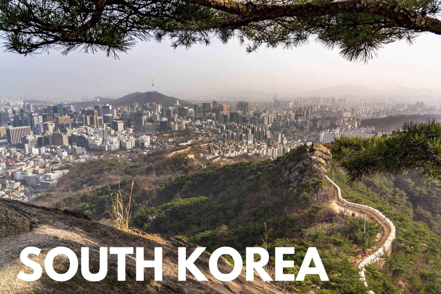 Photo of the city of Seoul South Korea behind hills with a stone wall and a tree branch, and the words South Korea overlaid
