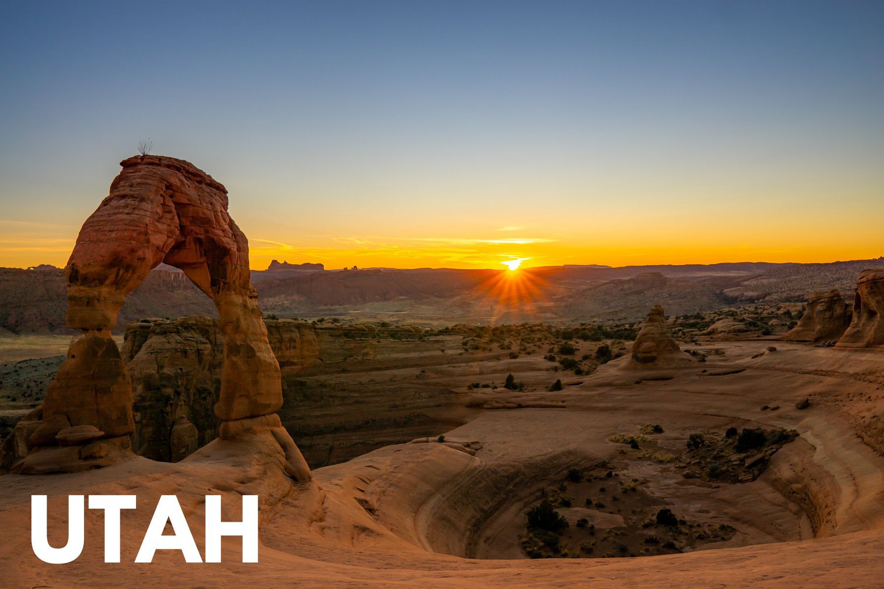 Photo of Delicate Arch and a bowl shaped depression with the sun setting on the distant red rock horizon and the word Utah overlaid