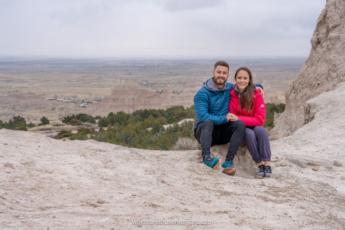 Couple sat together for a photo on a rocky ledge with a far reaching valley view behind on a cloudy day