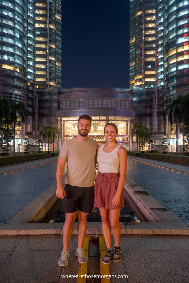 Couple standing next to each other in shorts and t-shirts for a photo at the base of twin skyscrapers Petronas Towers in Kuala Lumpur at night