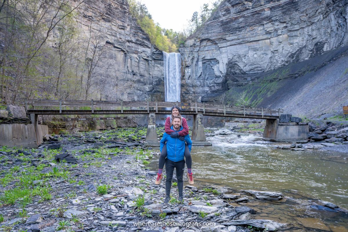 Hiker jumping up on the back of another hiker for a photo with a river, bridge and waterfall