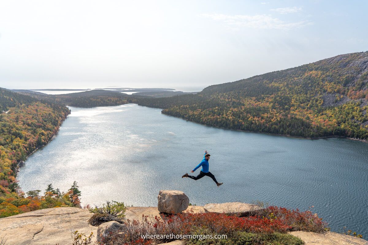 Photo taken from far away of a hiker jumping between rocks in front of a lake on a very sunny and hazy day