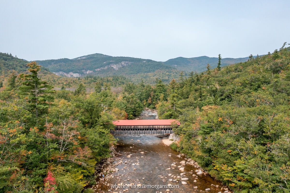 Drone photo from above of the Albany Covered Bridge with its red roof crossing a river and surrounded by trees and rolling hills on the Kancamagus Scenic Byway